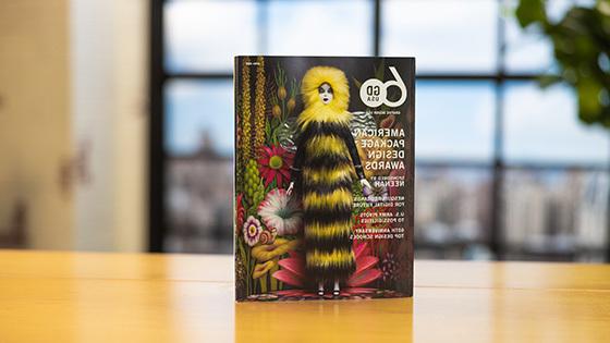 Graphic Design USA’s April 2023 issue sits upright on a wooden table. The cover features a female doll wearing a long furry black-and-yellow dress with a hood. She is standing in front of a psychedelic floral background.